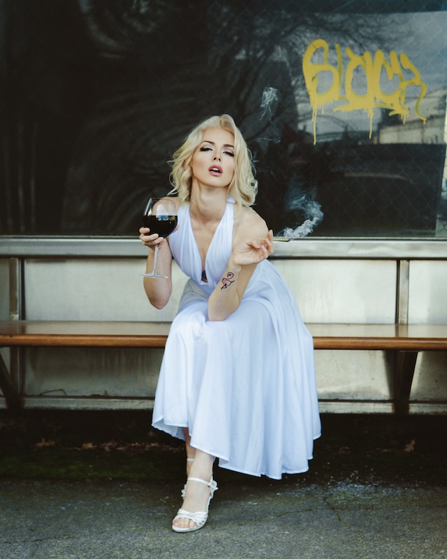 Woman in a white dress, smoking a cigarette while holding a glass of wine.
