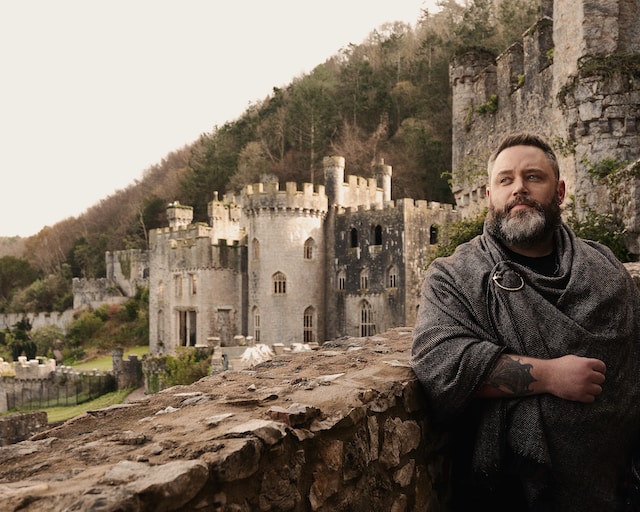 A man, garbed in what seemed like a recreation of medieval fashion, standing in front of a castle